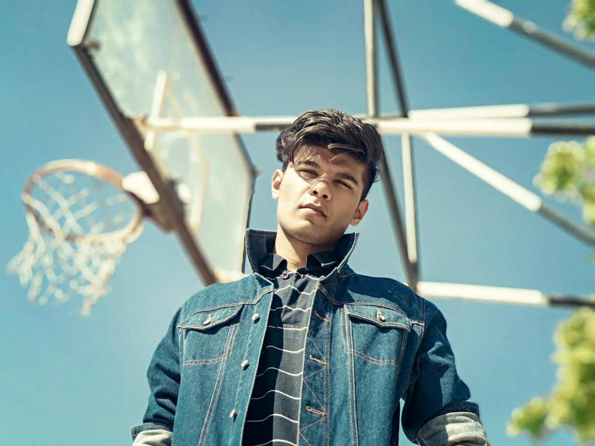 Teen boy in a denim jacket standing under a basketball hoop against a blue sky